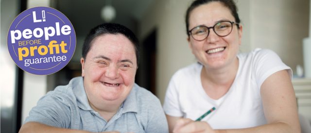 two people smiling at the camera. one is reading braille.