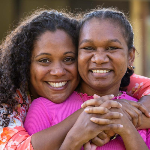 Two young aboriginal women outdoors with their arms around each other smiling at the camera.