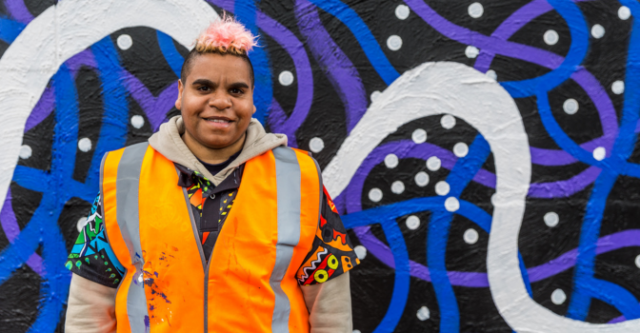 Jackie Saunders wearing a hi-vis vest, in front a wall with blue, purple and white swirls painted on it.