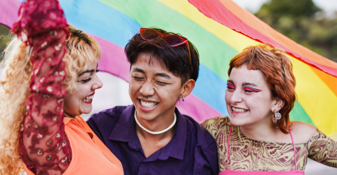 Three people embracing and laughing holding a pride flag above their head.