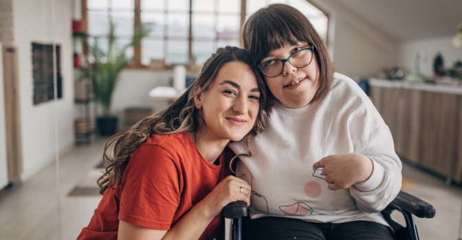 Young girl with disability and her carer, leaning in for a picture in a kitchen.