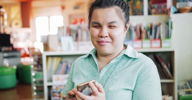 A woman with low vision is in a library spacce using her phone and smiling.