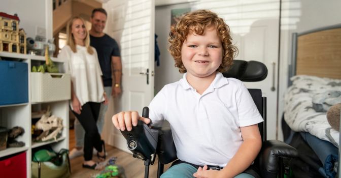 Cheerful young boy with muscular dystrophy in his bedroom, using powered wheelchair, mother and father smiling and watching in the background