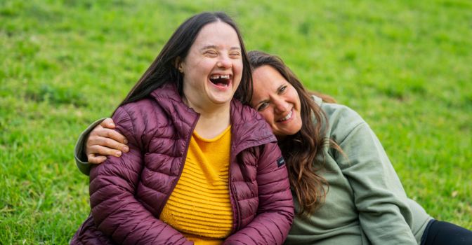 To women wearing winter jackets, sitting on the grass laughing.